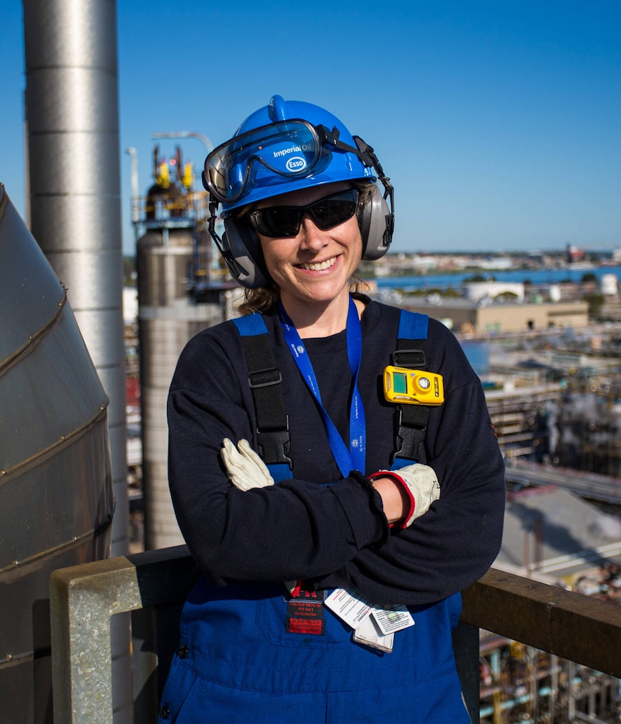 Profissional técnica sorrindo em planta industrial, com uniforme e capacete de segurança